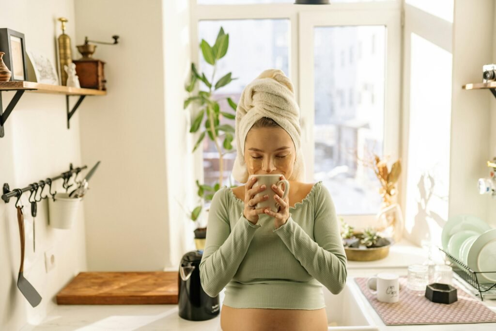 Pregnant woman with towel on head relaxes with tea in sunlight-filled kitchen. Cozy and serene atmosphere.